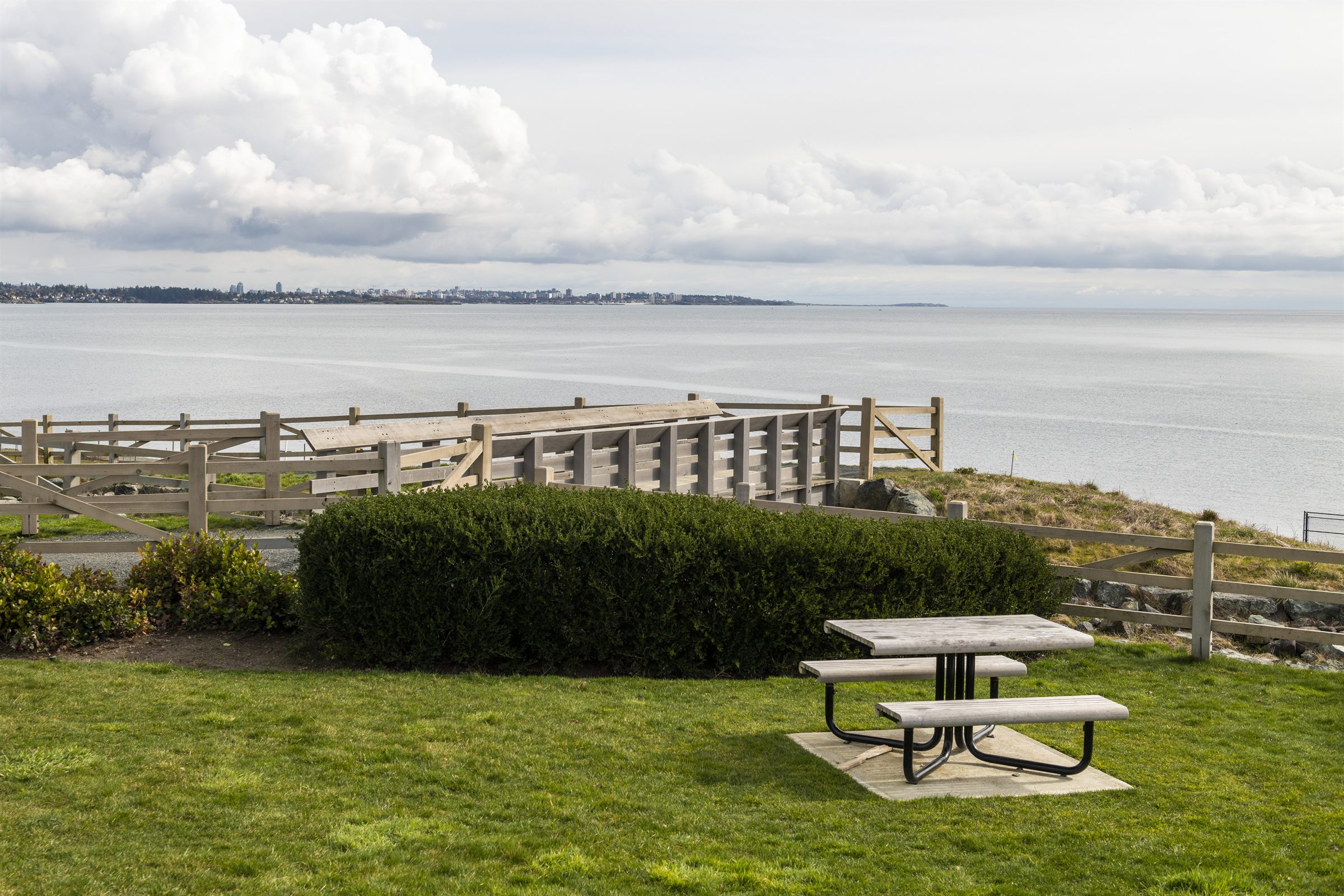 Waterfront park on the West Shore with a picnic table and split rail fence above the ocean, Victoria in the distance